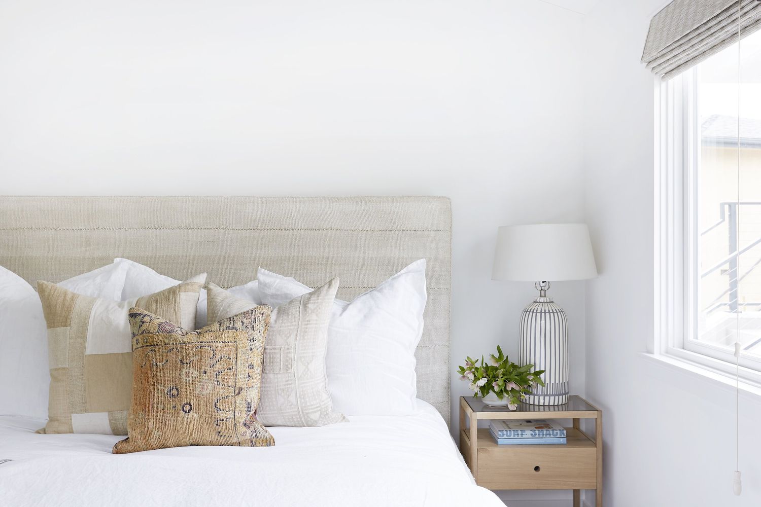 Un dormitorio lleno de blanco con un cabecero beige y varios cojines arrojadizos color almendra